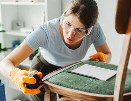 A woman in safety glasses measures a chair seat.